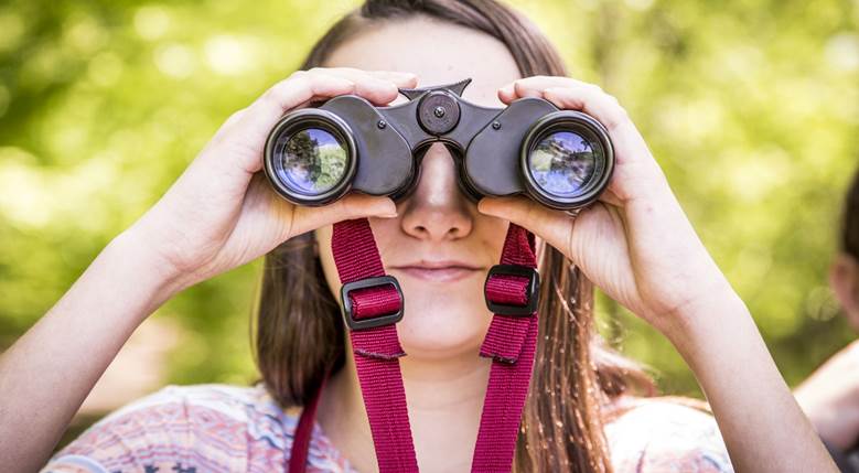A young girl looks through binoculars directly at the camera.